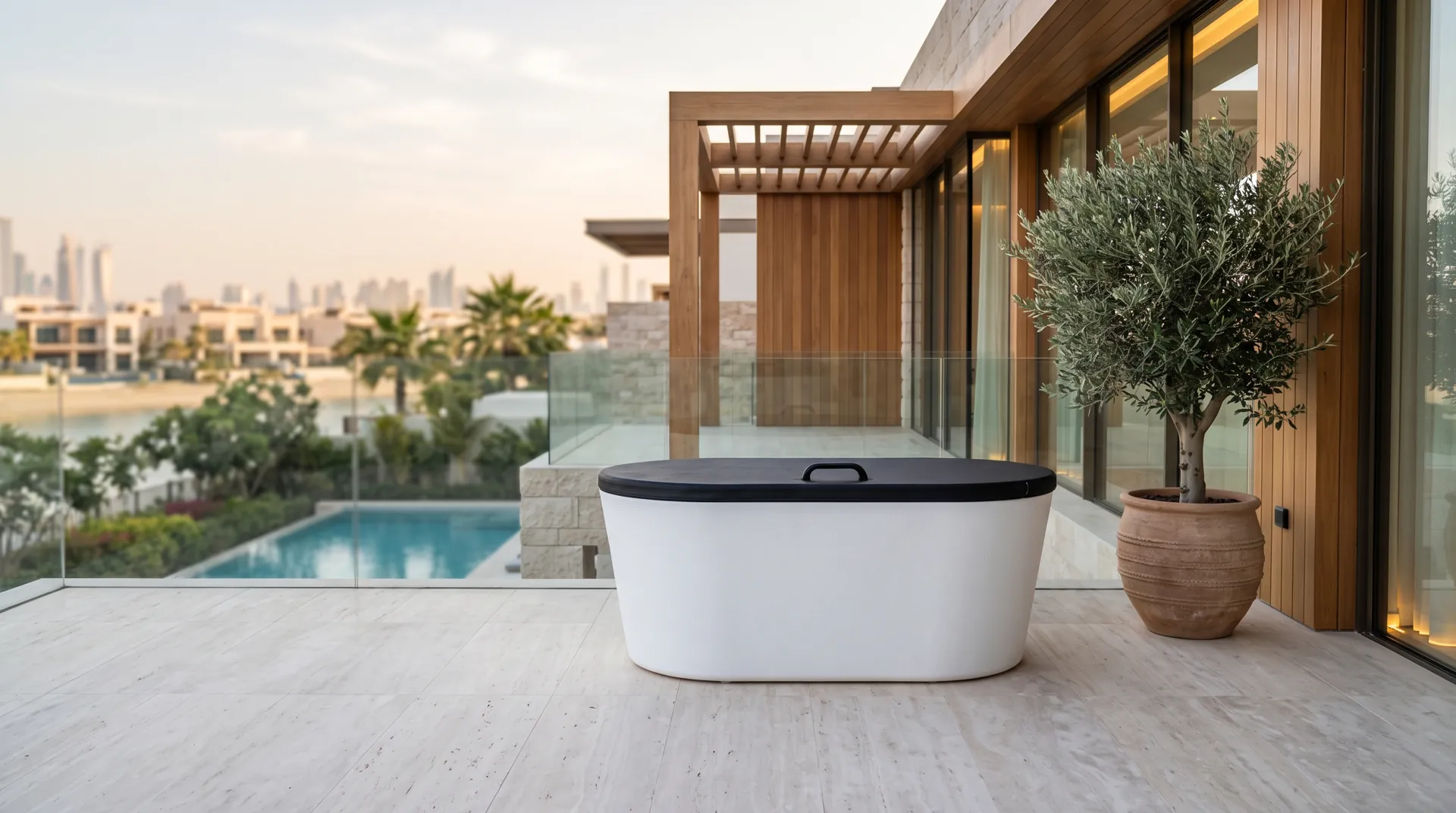 A white oval cold plunge tub with a black lid sits on a luxury terrace beside a potted olive tree overlooking a pool