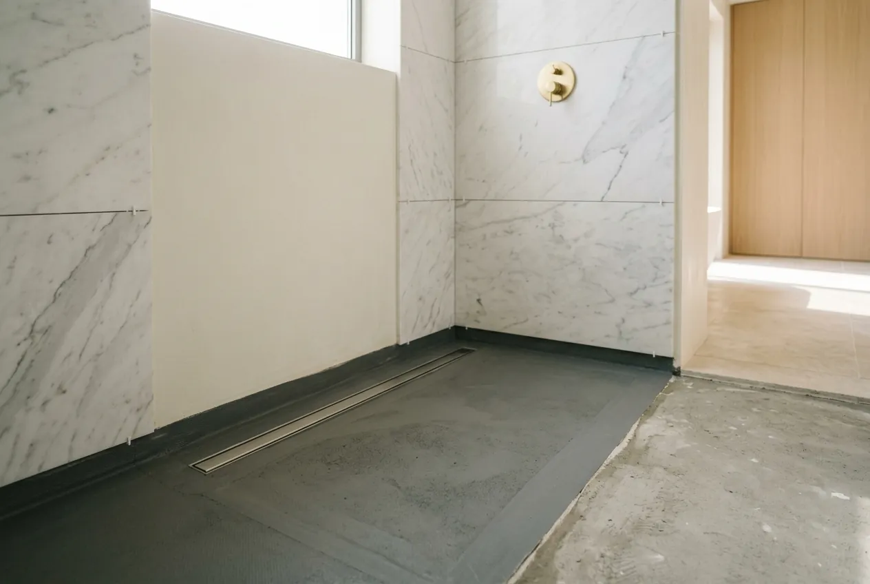 A walk-in shower under construction with waterproofed screed, a linear drain, and partially tiled marble walls
